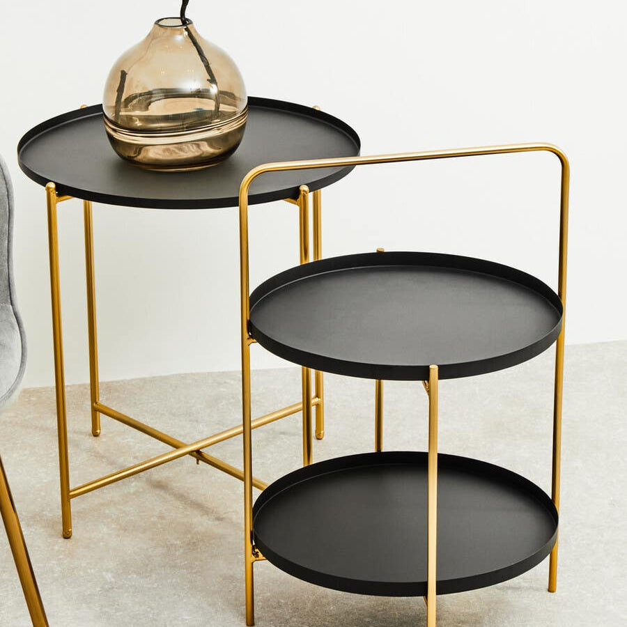 Black top side table with a round tray design and gold metal frame, featuring two tiers for storage or display. The upper tier is slightly smaller and holds a decorative vase, while the lower tier is larger and open. The table is set against a neutral background