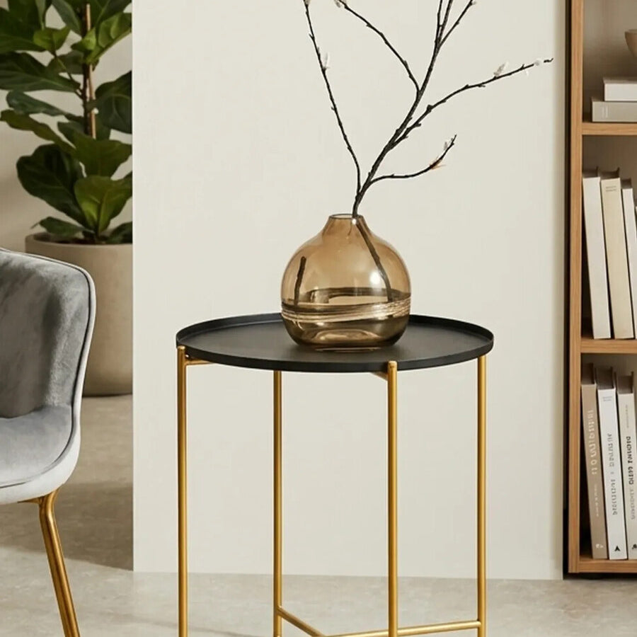 A black top side table with a round surface and gold metal legs, featuring a decorative vase with branches on top, set against a light-colored wall and a bookshelf in the background. A gray upholstered chair is partially visible to the left