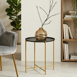 A black top side table with a round surface and gold metal legs, featuring a decorative vase with branches on top, positioned next to a light gray armchair and a wooden bookshelf in a modern living space