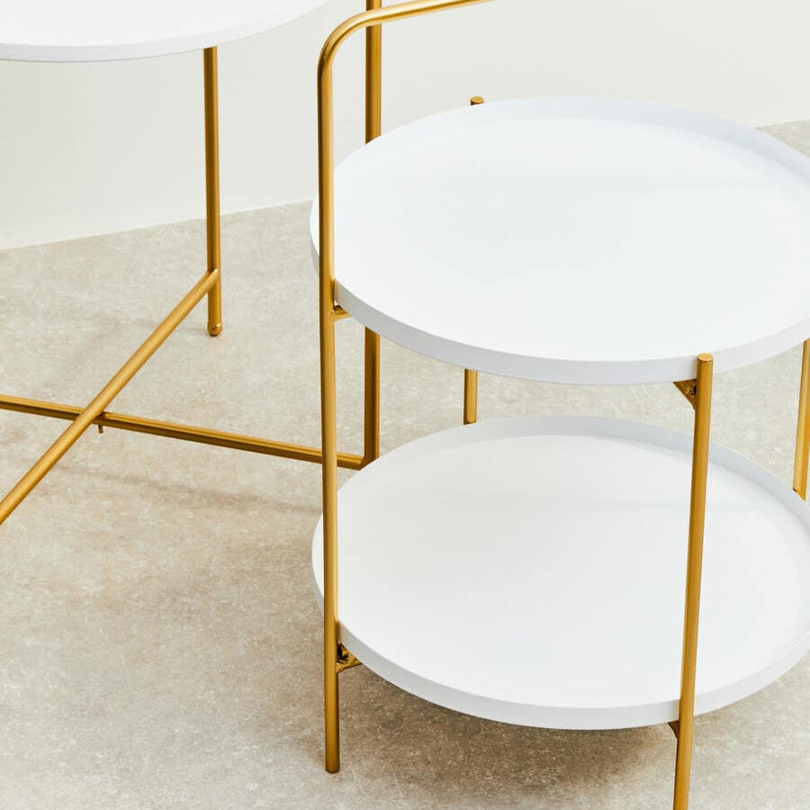 A modern side table featuring two round white tabletops supported by a sleek gold metal frame, with a minimalist design that complements contemporary decor. The table is positioned on a light-colored floor, enhancing its elegant appearance