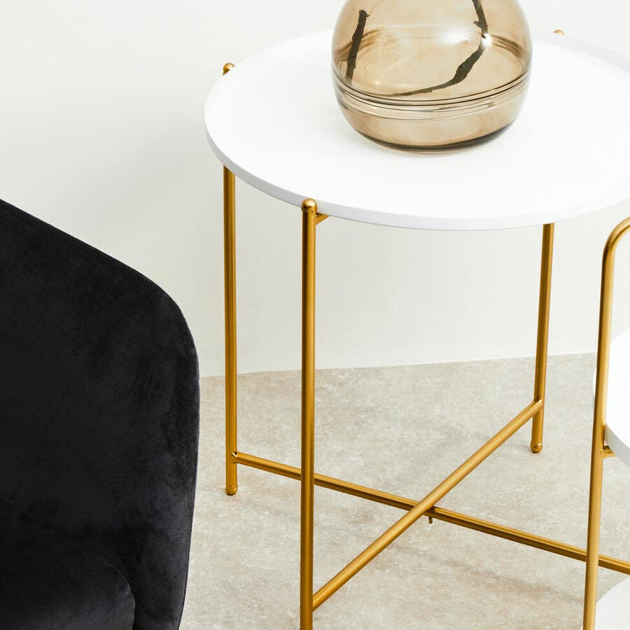 A white round side table with a smooth surface and gold metal legs, accompanied by a decorative glass vase on top. A black upholstered chair is partially visible beside the table, set against a light-colored floor