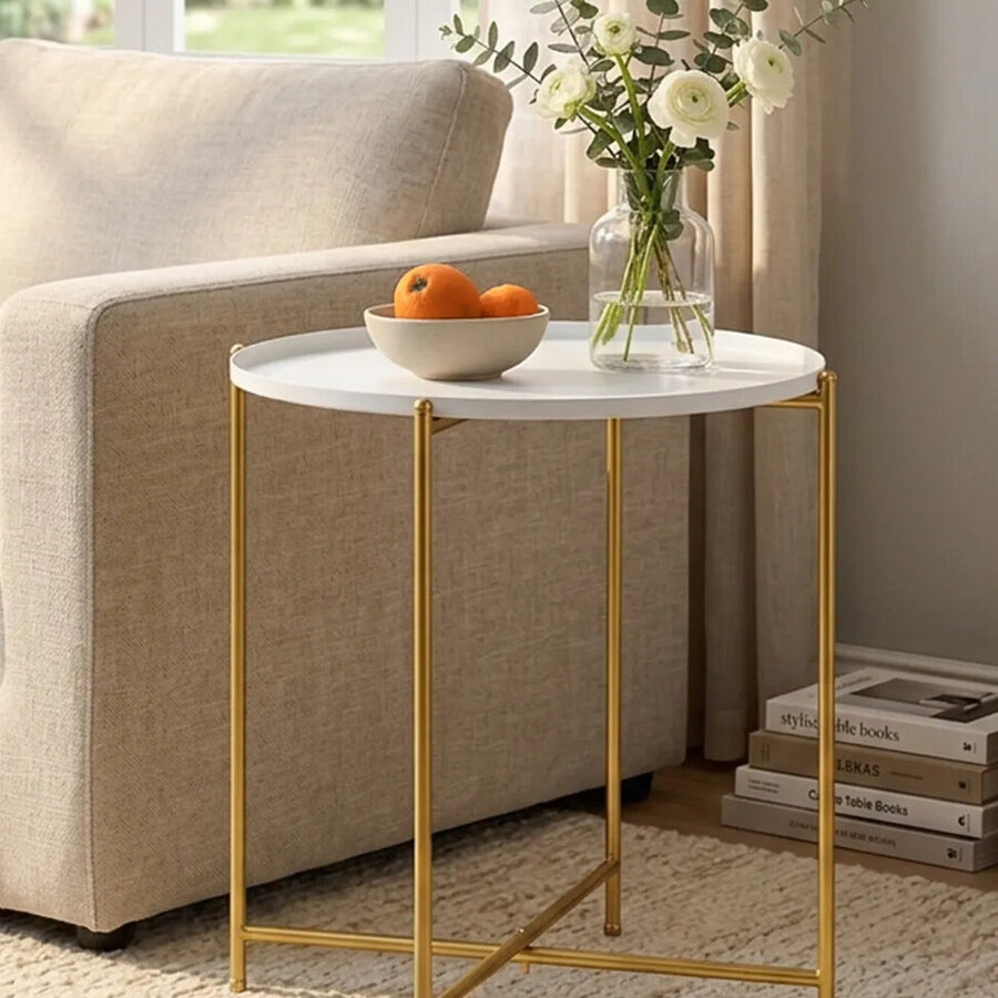 A white round side table with a smooth marble-like top and sleek gold metal legs, positioned next to a beige sofa. A bowl of oranges sits on the table, accompanied by a small vase with flowers. The setting features natural light coming through a nearby window, with a soft rug underneath