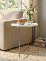 A white round side table with a smooth marble-like top and sleek gold metal legs, positioned next to a beige sofa. A bowl of oranges sits on the table, accompanied by a small vase with flowers. The setting features natural light coming through a nearby window, with a soft rug underneath