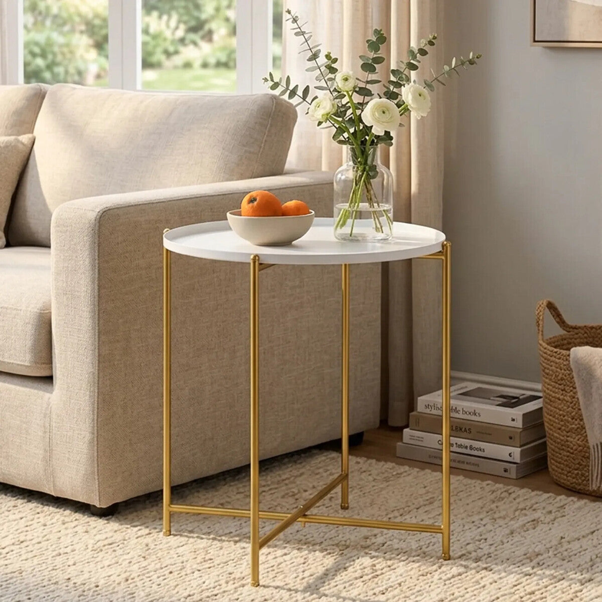 Round side table with a white marble top and gold metal legs, placed next to a beige sofa. A bowl of oranges sits on the table, and a vase with flowers is visible in the background. The setting features a light-colored rug and natural light coming through a window