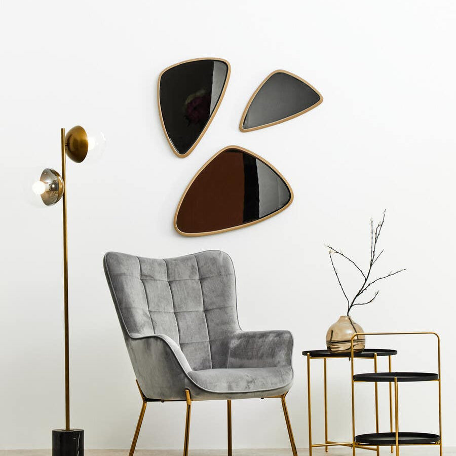 A stylish two-tier side table featuring a black top and gold metal frame, positioned next to a gray upholstered armchair. The table holds a decorative vase with a branch, and is complemented by three abstract wall mirrors in varying shapes and colors. A modern floor lamp stands nearby, enhancing the contemporary decor
