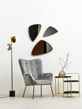 A stylish two-tier side table featuring a black top and gold metal frame, positioned next to a gray upholstered armchair. The table holds a decorative vase with a branch, and is complemented by three abstract wall mirrors in varying shapes and colors. A modern floor lamp stands nearby, enhancing the contemporary decor