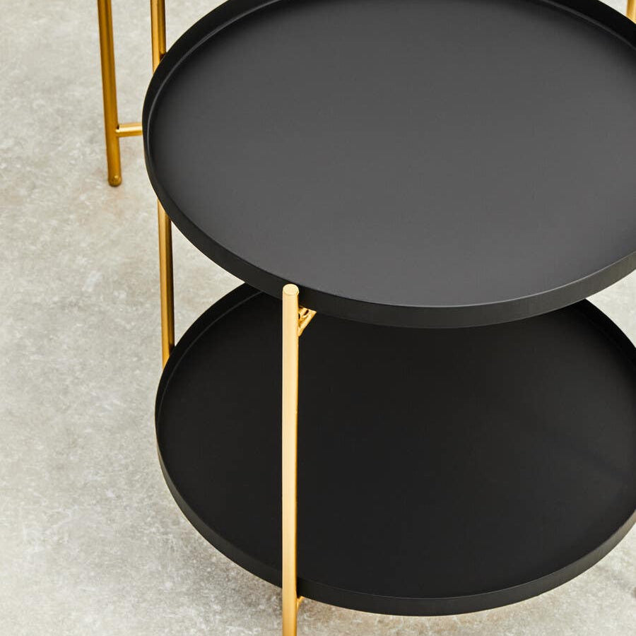 Two-tier side table featuring a sleek black circular top and bottom, supported by elegant gold metal legs. The design is modern and minimalist, suitable for various home decor styles. The surface is smooth and matte, contrasting with the shiny gold accents