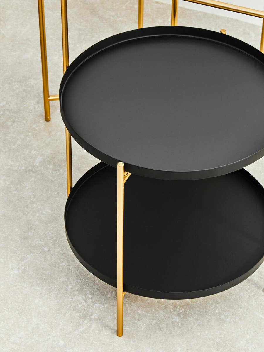 Two-tier side table featuring a sleek black circular top and bottom, supported by elegant gold metal legs. The design is modern and minimalist, suitable for various home decor styles. The surface is smooth and matte, contrasting with the shiny gold accents