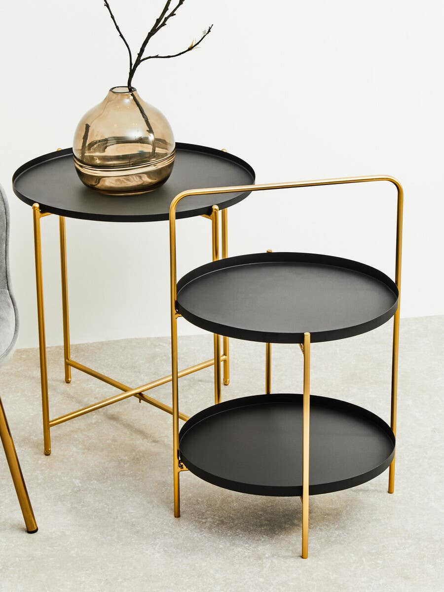 Two-tier side table featuring a sleek black finish with gold metal accents. The table has a circular top and a lower shelf, both in black, supported by a gold frame with a modern design. A decorative vase with a natural element is placed on the upper tier. The background is a neutral color, enhancing the table's elegant appearance