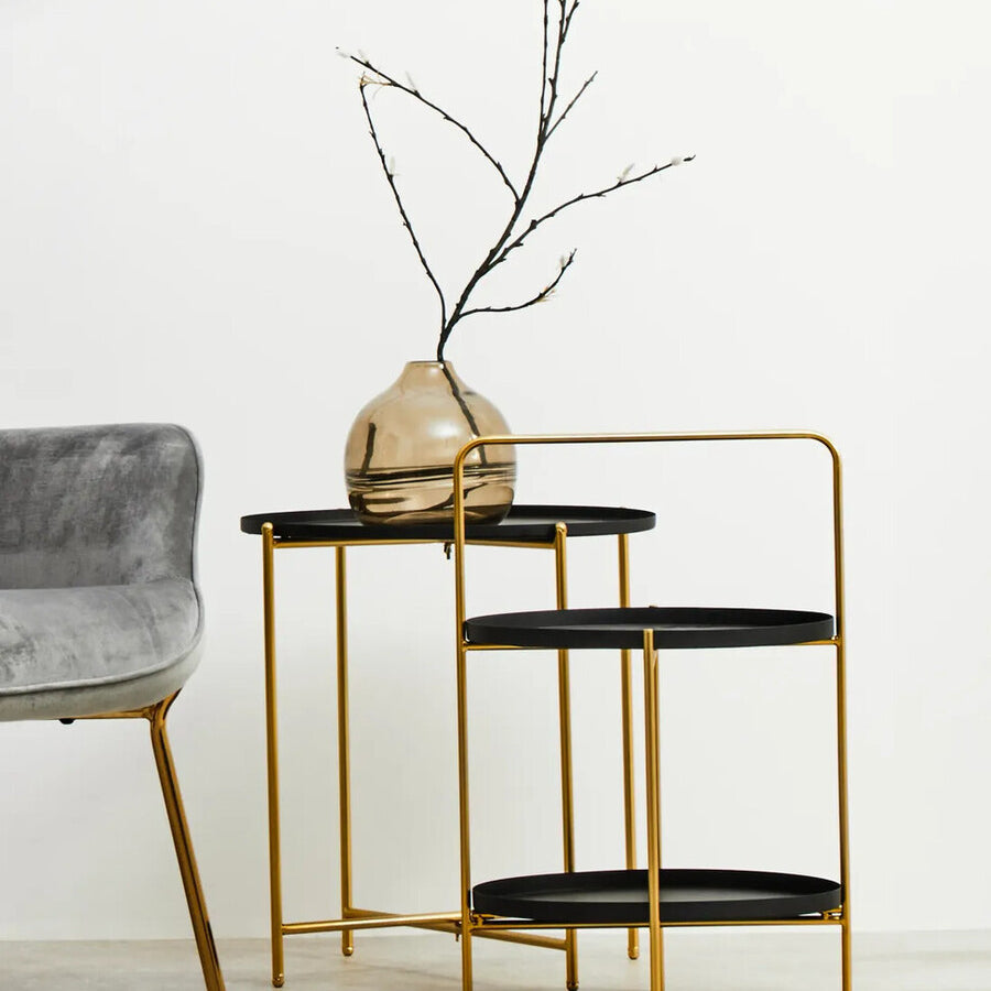 A two-tier side table featuring a sleek black finish on both tiers and a stylish gold metal frame. The top tier holds a decorative vase with a slender branch, while the bottom tier remains empty. The table is positioned next to a plush gray chair against a light-colored wall