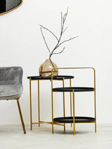 A two-tier side table featuring a sleek black finish on both tiers and a stylish gold metal frame. The top tier holds a decorative vase with a slender branch, while the bottom tier remains empty. The table is positioned next to a plush gray chair against a light-colored wall