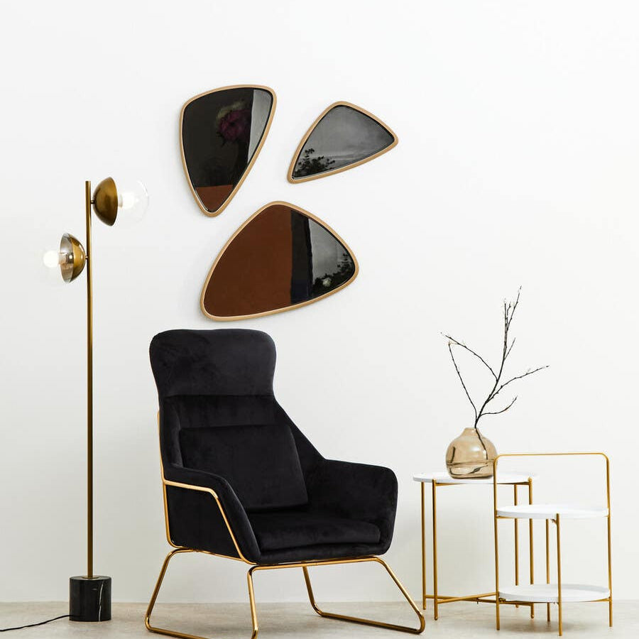 A stylish two-tier side table featuring a white top and gold metal frame, positioned next to a black upholstered armchair. The table is complemented by decorative elements, including a slender vase with branches and modern wall mirrors in abstract shapes above. A floor lamp with a gold finish stands nearby, enhancing the contemporary aesthetic of the space