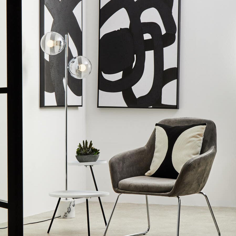 A modern side table featuring a white and black design, positioned next to a stylish chair with patterned cushions. The table has a sleek, minimalist frame and is complemented by abstract black and white wall art in the background. A small green plant adds a touch of color to the scene