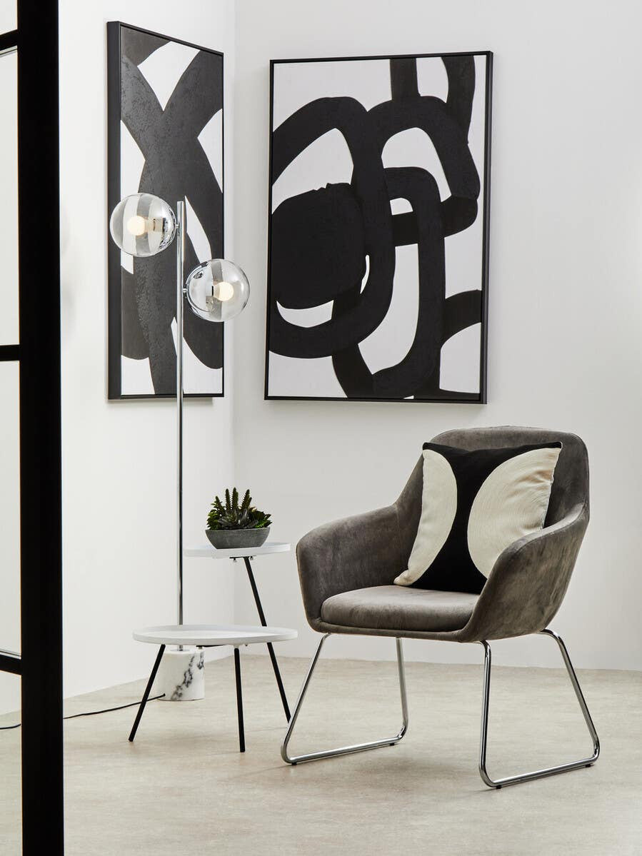A modern side table featuring a white and black design, positioned next to a stylish chair with patterned cushions. The table has a sleek, minimalist frame and is complemented by abstract black and white wall art in the background. A small green plant adds a touch of color to the scene