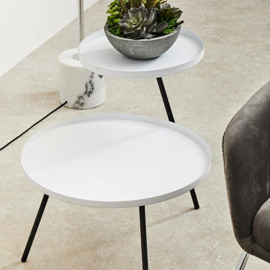 A modern side table featuring a round white top and slender black legs, with a smaller circular shelf positioned above the main surface. A decorative gray pot with a green plant sits on the top surface, and the table is placed on a light-colored floor