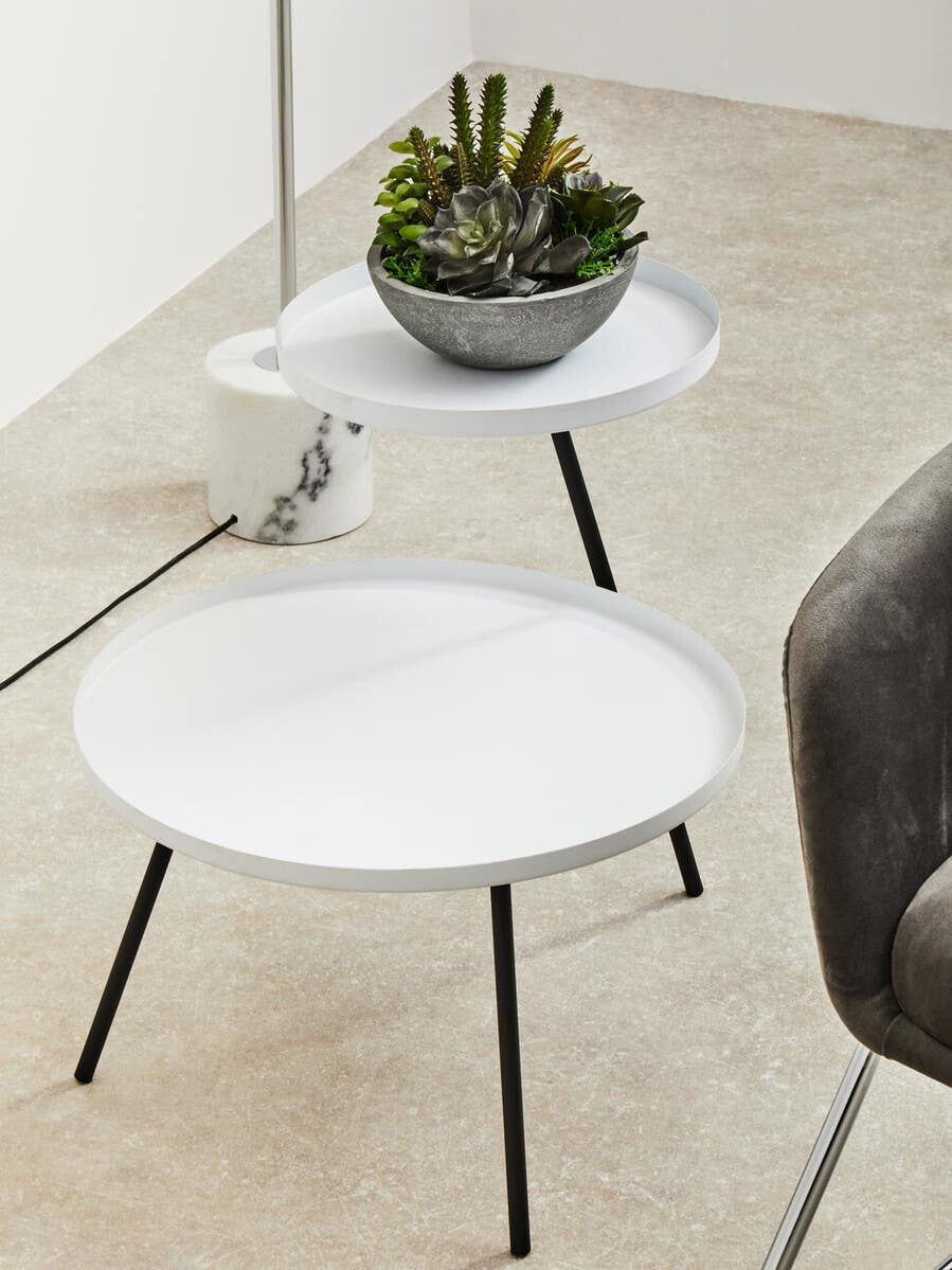 A modern side table featuring a round white top and slender black legs, with a smaller circular shelf positioned above the main surface. A decorative gray pot with a green plant sits on the top surface, and the table is placed on a light-colored floor