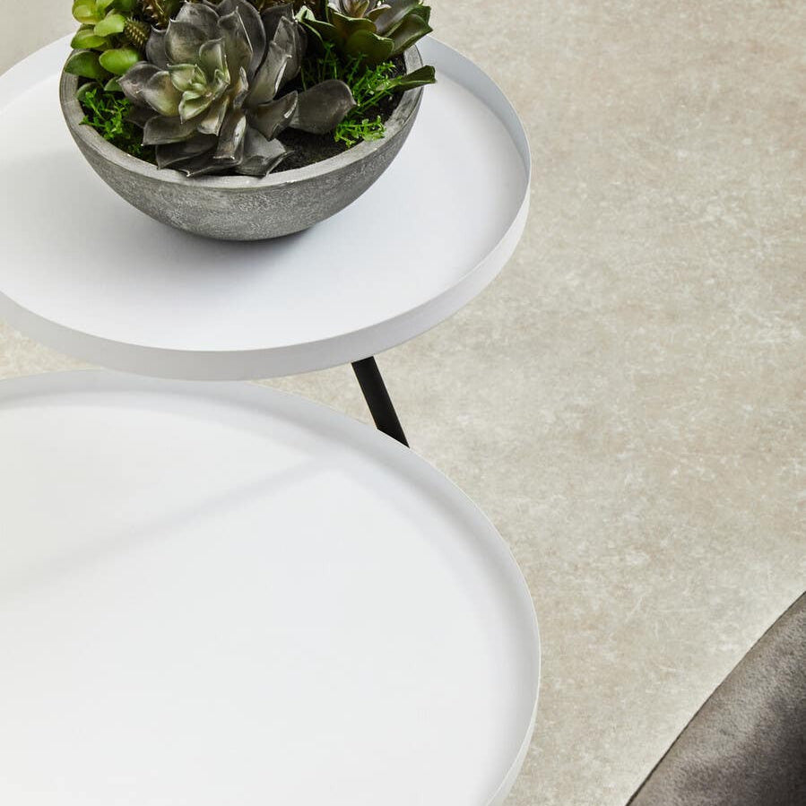 A modern side table featuring two circular tiers, one in white and the other in black, with a sleek design. A small potted plant sits on the upper tier, adding a touch of greenery. The table is positioned on a light-colored surface, highlighting its contemporary aesthetic