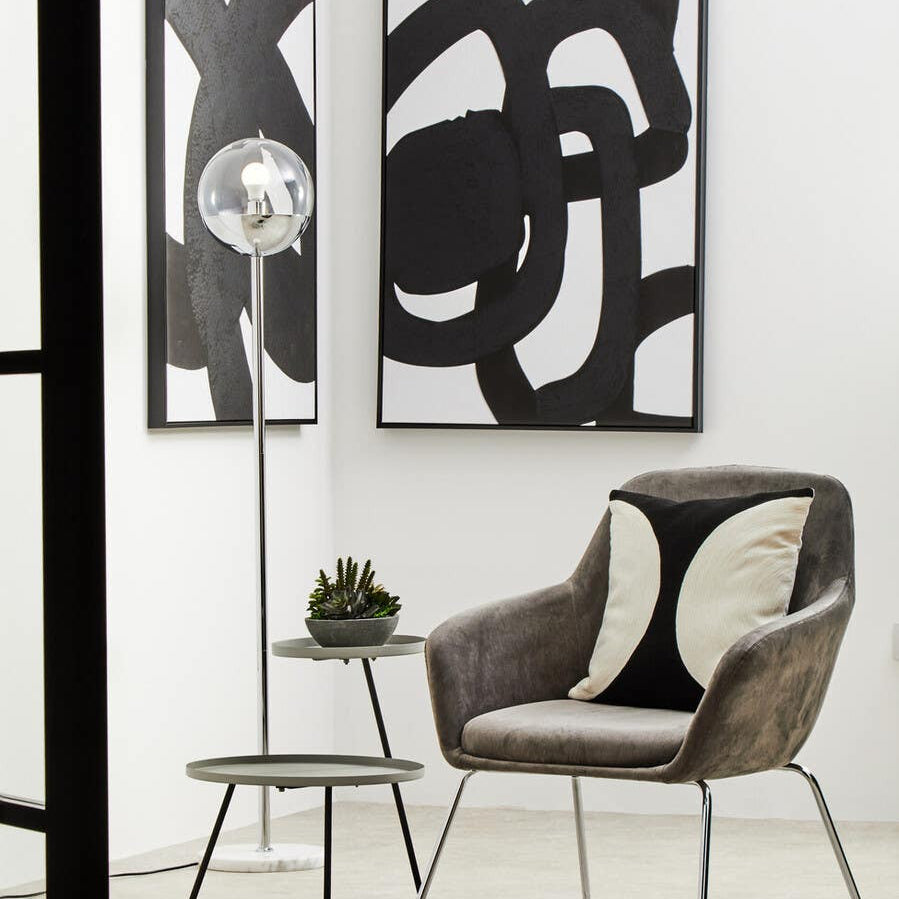 A modern side table featuring two shelves in a grey and black color scheme, positioned next to a stylish chair. The table has a sleek design with clean lines, complementing the abstract wall art in the background. A small decorative plant sits on the top shelf, enhancing the contemporary aesthetic of the room