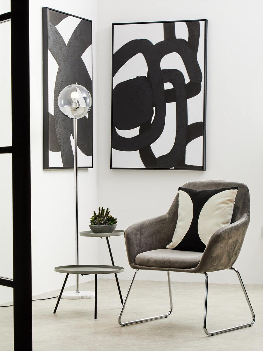 A modern side table featuring two shelves in a grey and black color scheme, positioned next to a stylish chair. The table has a sleek design with clean lines, complementing the abstract wall art in the background. A small decorative plant sits on the top shelf, enhancing the contemporary aesthetic of the room