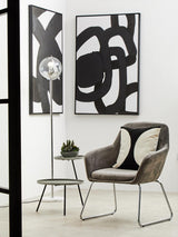 A modern side table featuring two shelves in a grey and black color scheme, positioned next to a stylish chair. The table has a sleek design with clean lines, complementing the abstract wall art in the background. A small decorative plant sits on the top shelf, enhancing the contemporary aesthetic of the room