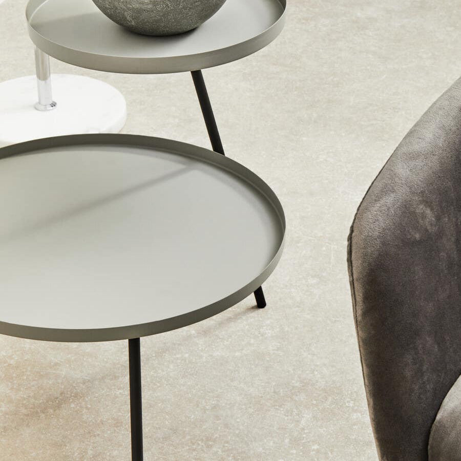 A modern two-shelf side table featuring a grey top and black metal legs, with a round design. The upper shelf holds a small decorative bowl with a plant, while the lower shelf is empty. The table is positioned on a light-colored carpet, next to a plush grey chair