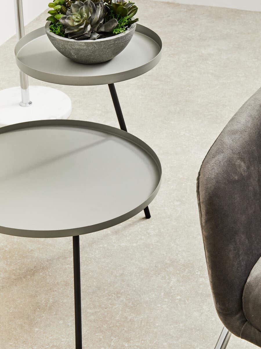 A modern two-shelf side table featuring a grey top and black metal legs, with a round design. The upper shelf holds a small decorative bowl with a plant, while the lower shelf is empty. The table is positioned on a light-colored carpet, next to a plush grey chair