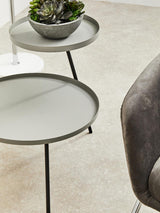 A modern two-shelf side table featuring a grey top and black metal legs, with a round design. The upper shelf holds a small decorative bowl with a plant, while the lower shelf is empty. The table is positioned on a light-colored carpet, next to a plush grey chair