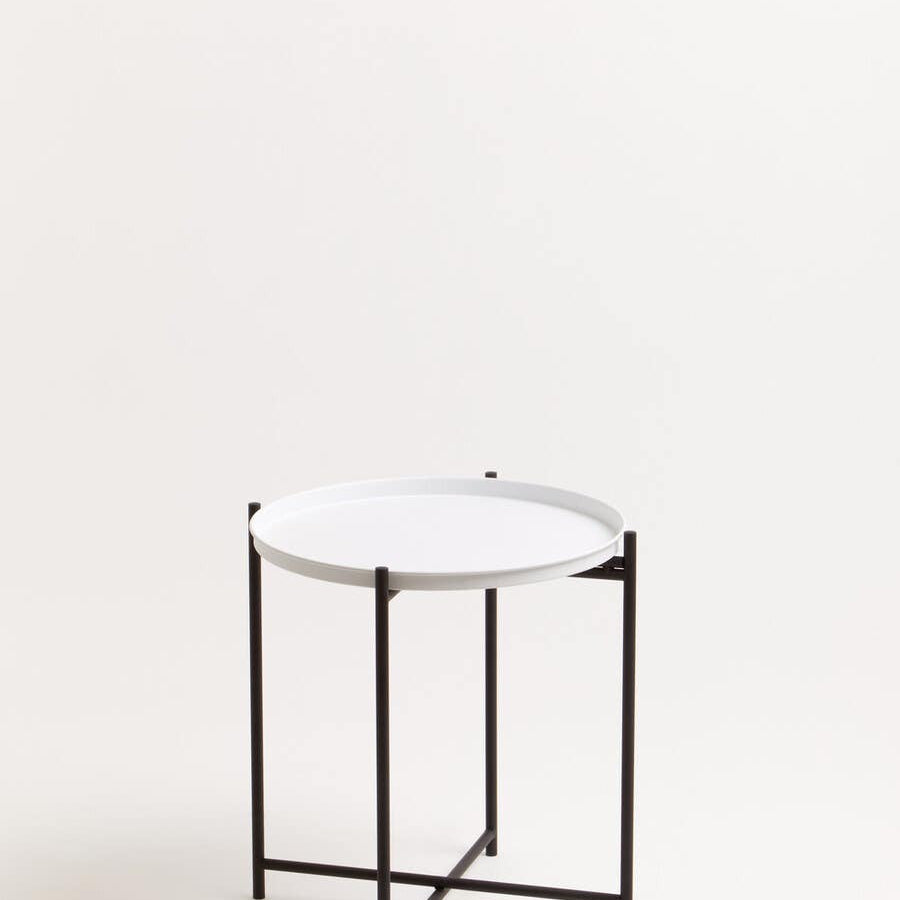 Round white tabletop supported by a sleek black metal frame, featuring a minimalist design with four legs and a crossbar for stability. The background is a neutral color, enhancing the modern aesthetic of the side table