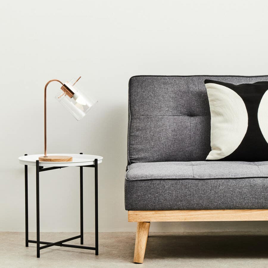 A round white top side table with a wooden base and metal legs, positioned next to a gray sofa adorned with black and white decorative pillows. A modern lamp with a curved neck is placed on the table, illuminating the space. The background features a light-colored wall and a neutral-toned floor