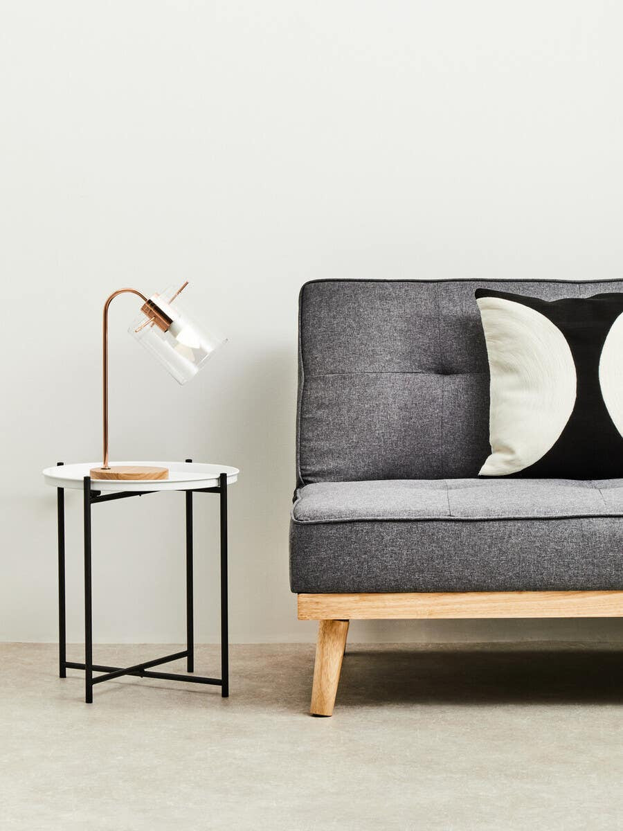 A round white top side table with a wooden base and metal legs, positioned next to a gray sofa adorned with black and white decorative pillows. A modern lamp with a curved neck is placed on the table, illuminating the space. The background features a light-colored wall and a neutral-toned floor