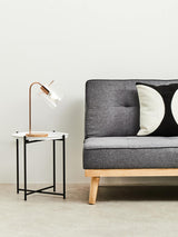 A round white top side table with a wooden base and metal legs, positioned next to a gray sofa adorned with black and white decorative pillows. A modern lamp with a curved neck is placed on the table, illuminating the space. The background features a light-colored wall and a neutral-toned floor