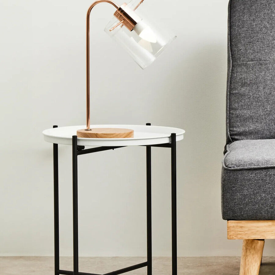 Round white top side table with a sleek black metal frame, positioned next to a gray upholstered armchair. A modern lamp with a curved neck and wooden base is placed on the table, providing a stylish accent to the setup. The background features a light-colored wall, enhancing the contemporary look of the furniture arrangement