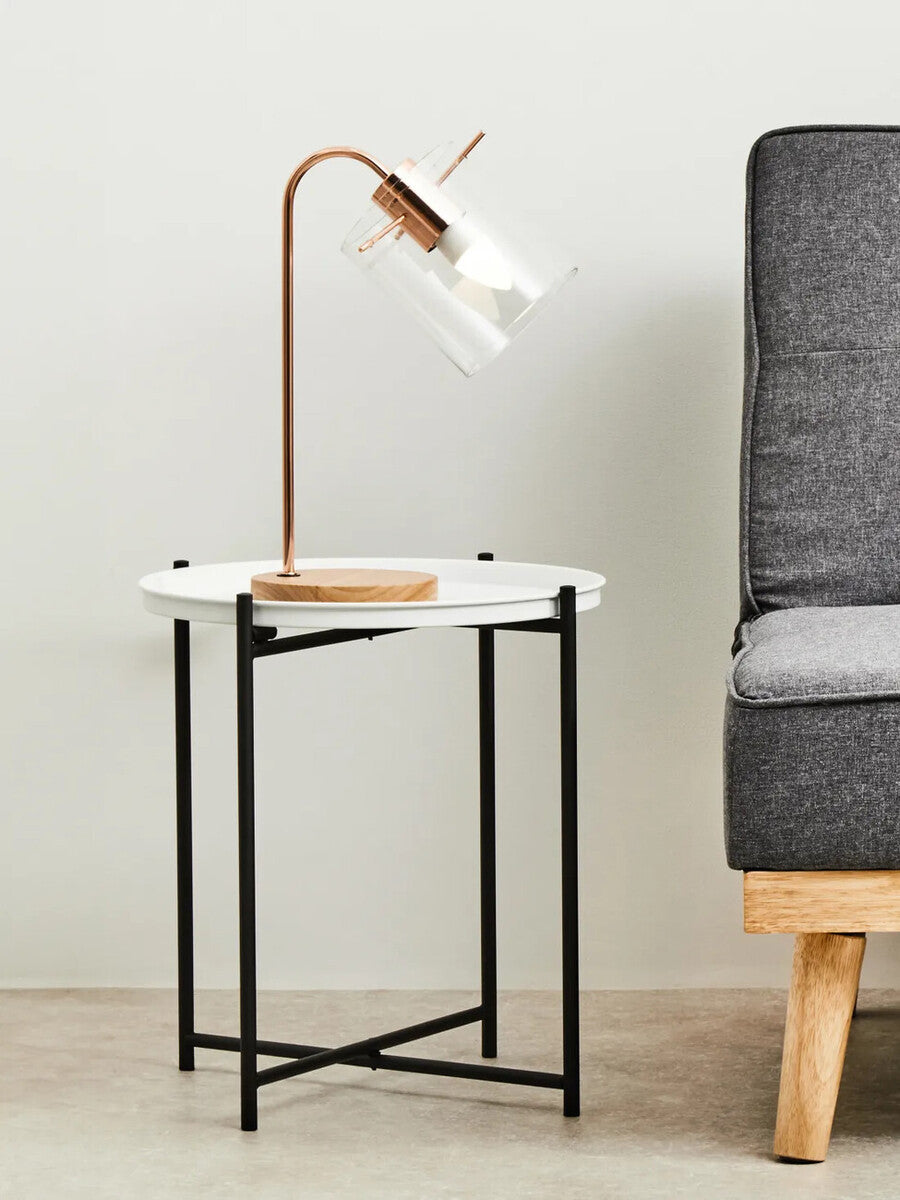 Round white top side table with a sleek black metal frame, positioned next to a gray upholstered armchair. A modern lamp with a curved neck and wooden base is placed on the table, providing a stylish accent to the setup. The background features a light-colored wall, enhancing the contemporary look of the furniture arrangement