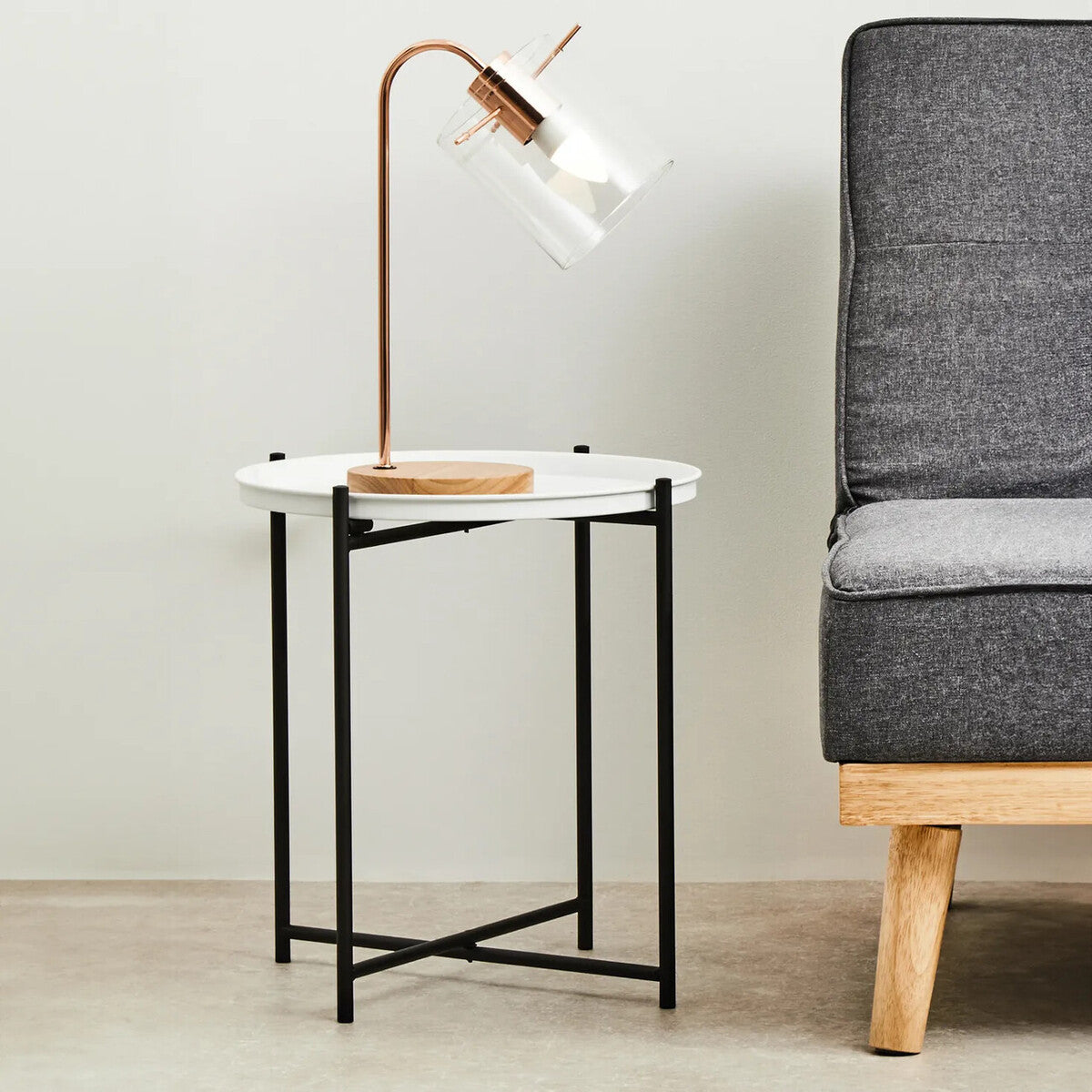 Round white top side table with a black metal frame, featuring a minimalist design. A stylish lamp with a copper finish is placed on the table, next to a gray upholstered sofa with wooden legs. The background is a light-colored wall and floor, creating a modern living space ambiance