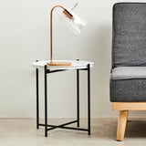 Round white top side table with a black metal frame, featuring a minimalist design. A stylish lamp with a copper finish is placed on the table, next to a gray upholstered sofa with wooden legs. The background is a light-colored wall and floor, creating a modern living space ambiance