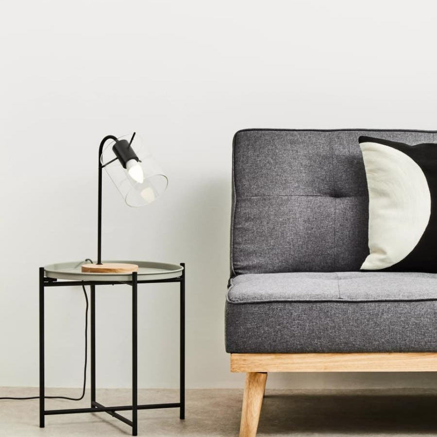 Round grey top side table with a black metal frame and wooden legs, positioned next to a modern grey sofa with tufted upholstery and a black and white decorative pillow. A sleek black lamp with a white shade is placed on the table, and the background features a plain white wall and light-colored flooring