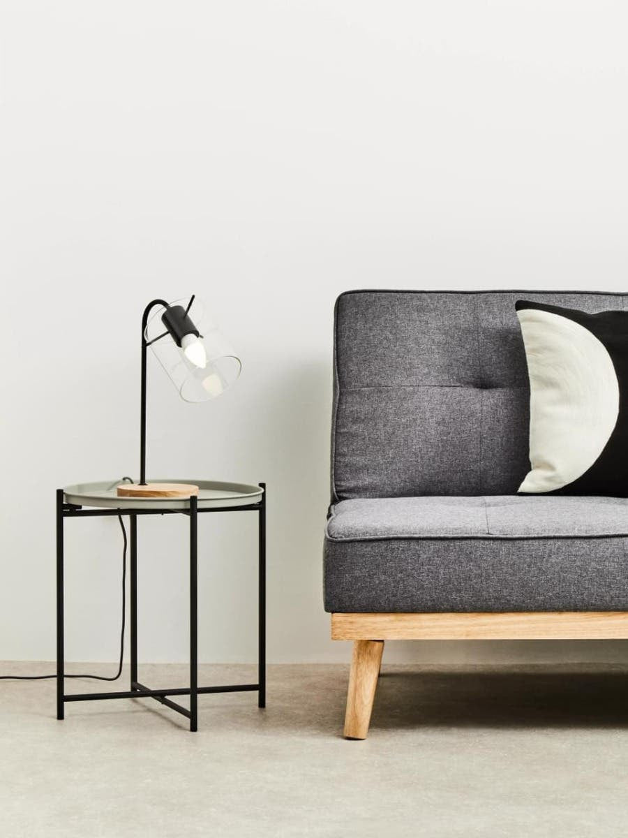 Round grey top side table with a black metal frame and wooden legs, positioned next to a modern grey sofa with tufted upholstery and a black and white decorative pillow. A sleek black lamp with a white shade is placed on the table, and the background features a plain white wall and light-colored flooring