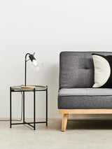 Round grey top side table with a black metal frame and wooden legs, positioned next to a modern grey sofa with tufted upholstery and a black and white decorative pillow. A sleek black lamp with a white shade is placed on the table, and the background features a plain white wall and light-colored flooring