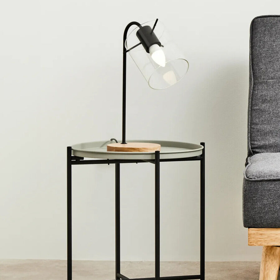 Round grey top side table with a black metal frame, featuring a wooden accent on the side. A stylish lamp with a clear bulb is positioned on the table, and a grey sofa is visible in the background against a light-colored wall
