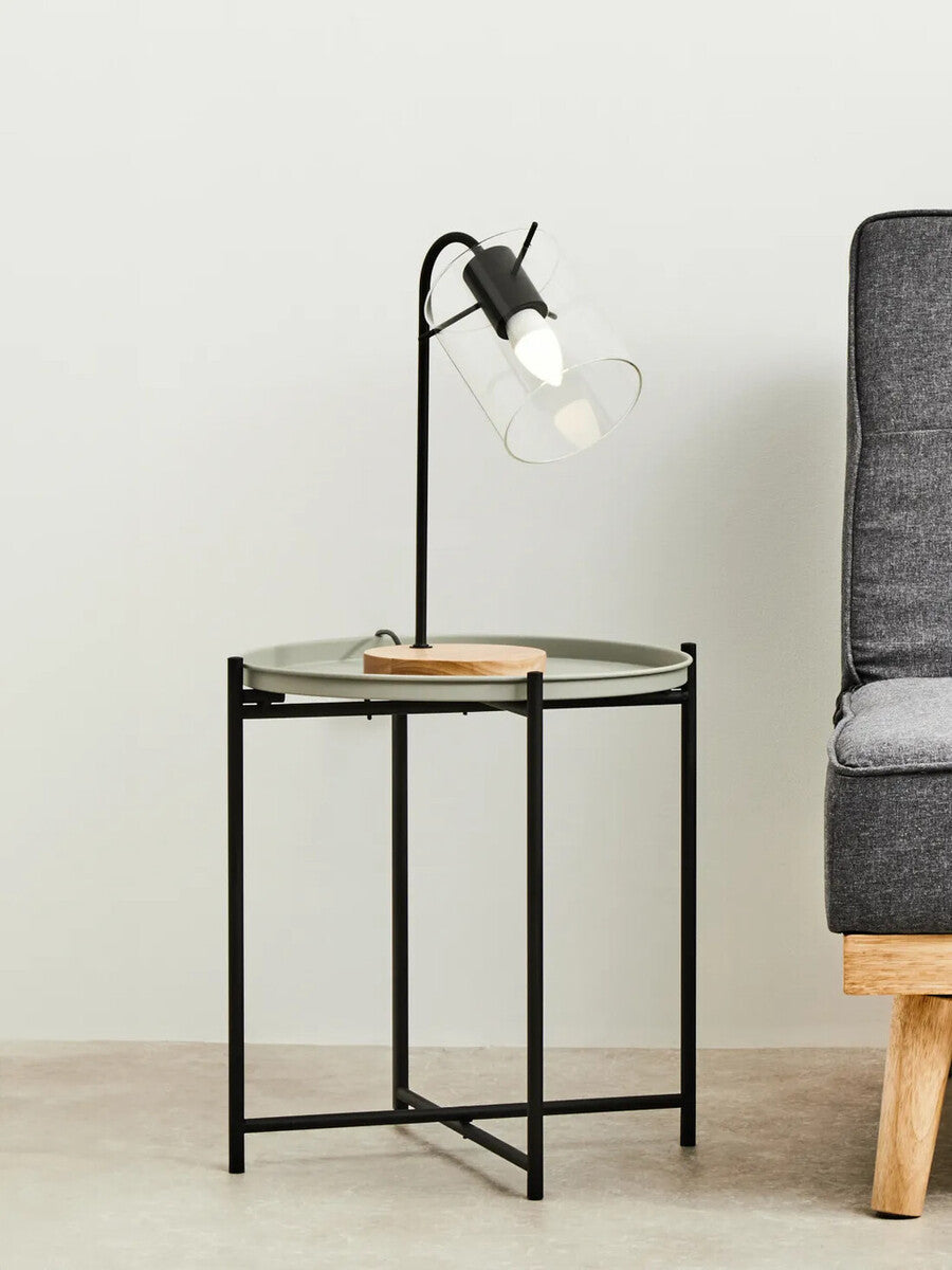 Round grey top side table with a black metal frame, featuring a wooden accent on the side. A stylish lamp with a clear bulb is positioned on the table, and a grey sofa is visible in the background against a light-colored wall