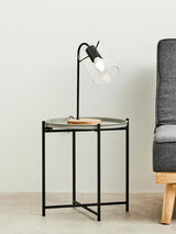 Round grey top side table with a black metal frame, featuring a wooden accent on the side. A stylish lamp with a clear bulb is positioned on the table, and a grey sofa is visible in the background against a light-colored wall