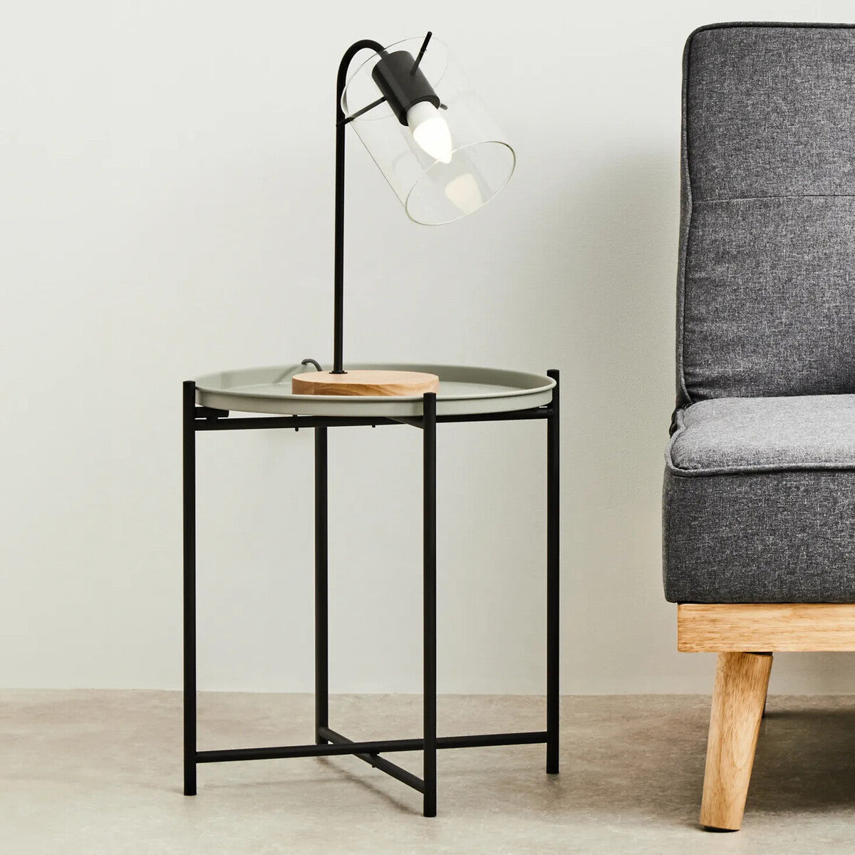 Round grey top side table with a black metal frame, featuring a wooden accent on the top. A modern lamp with a clear bulb is positioned on the table, and a grey upholstered sofa is visible in the background, along with light-colored flooring and a neutral wall