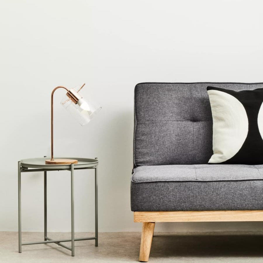 Grey iron side table with a minimalist design, featuring a sleek rectangular top and slender legs, positioned next to a modern grey sofa with tufted upholstery and decorative black and white pillows. A stylish lamp with a curved neck sits atop the table, illuminating the space against a light-colored wall and neutral floor