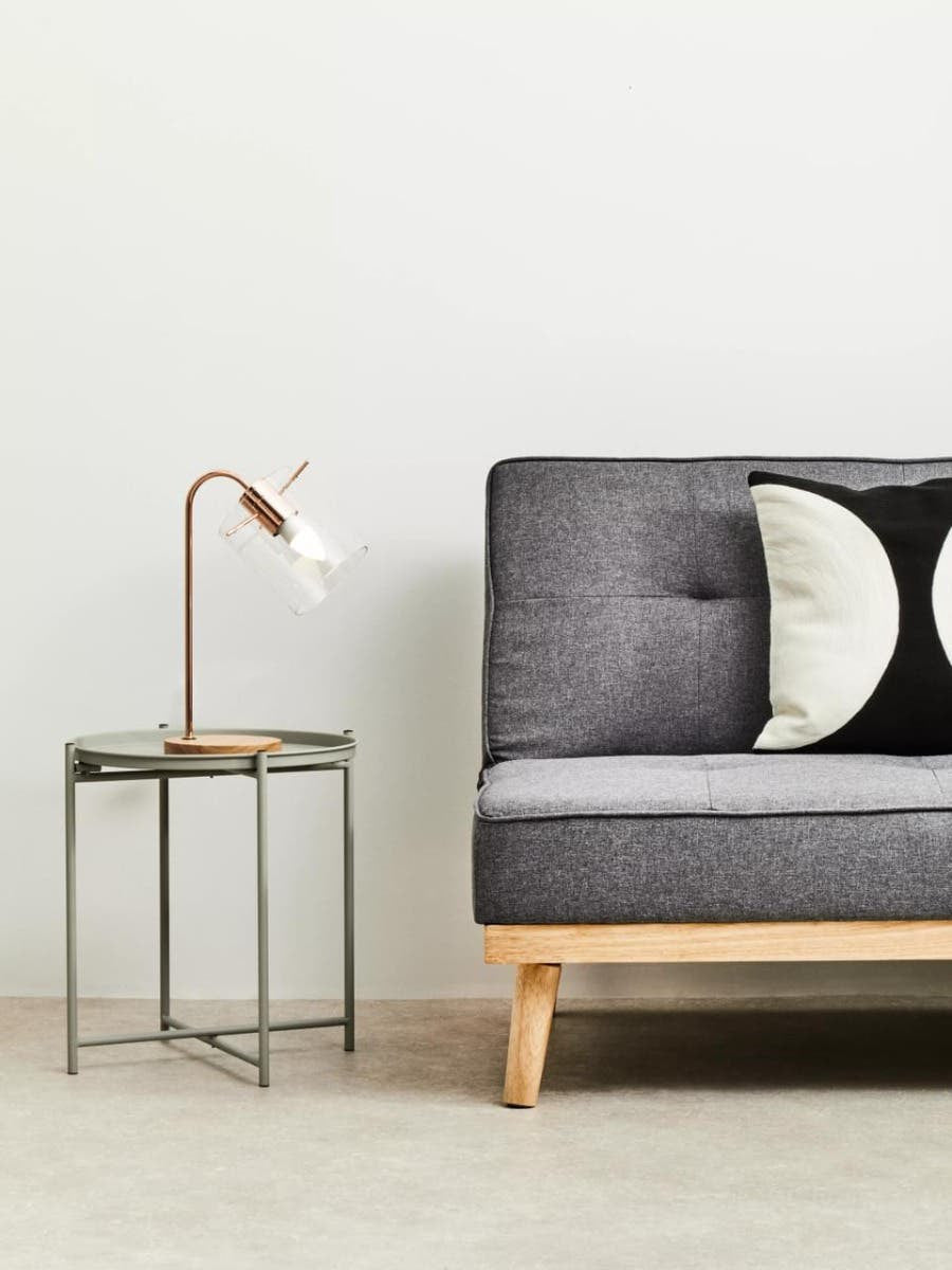 Grey iron side table with a minimalist design, featuring a sleek rectangular top and slender legs, positioned next to a modern grey sofa with tufted upholstery and decorative black and white pillows. A stylish lamp with a curved neck sits atop the table, illuminating the space against a light-colored wall and neutral floor