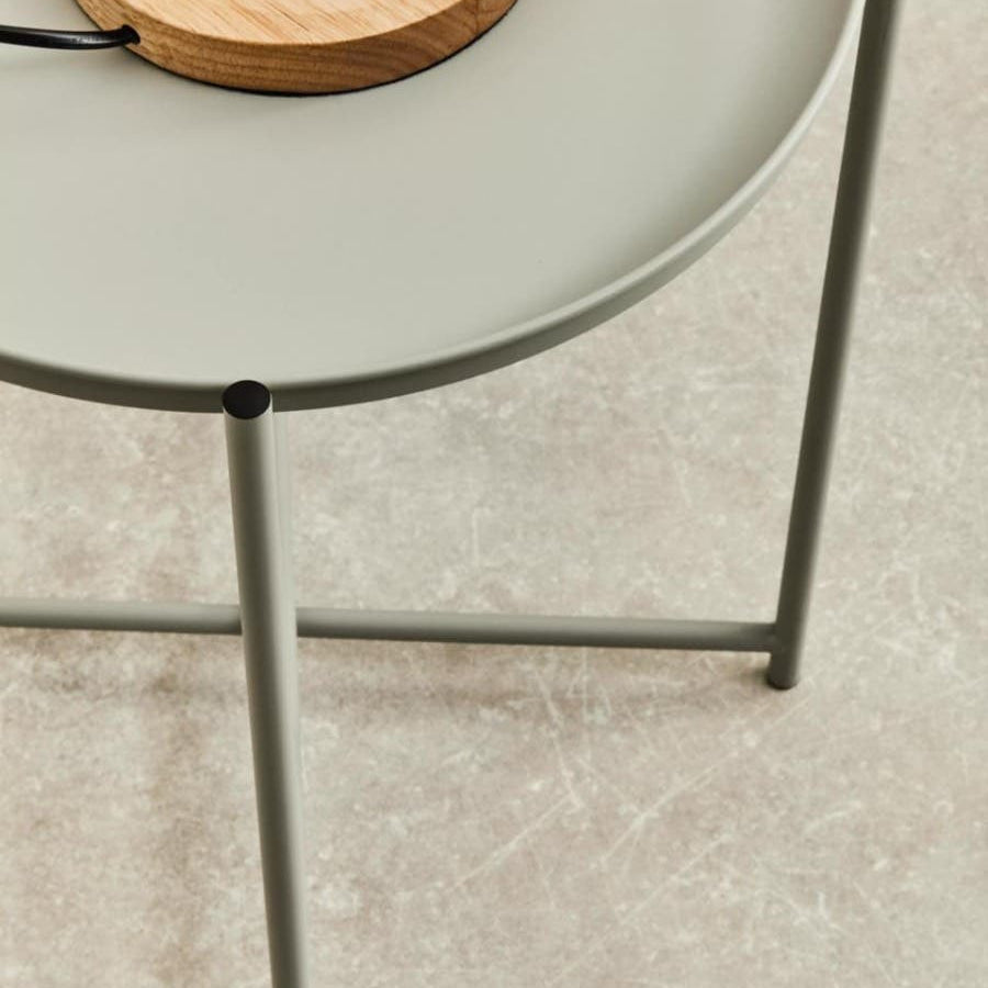 A grey iron side table with a round top and slender legs, featuring a wooden lamp base on the surface. The table is positioned on a light-colored floor, highlighting its modern design