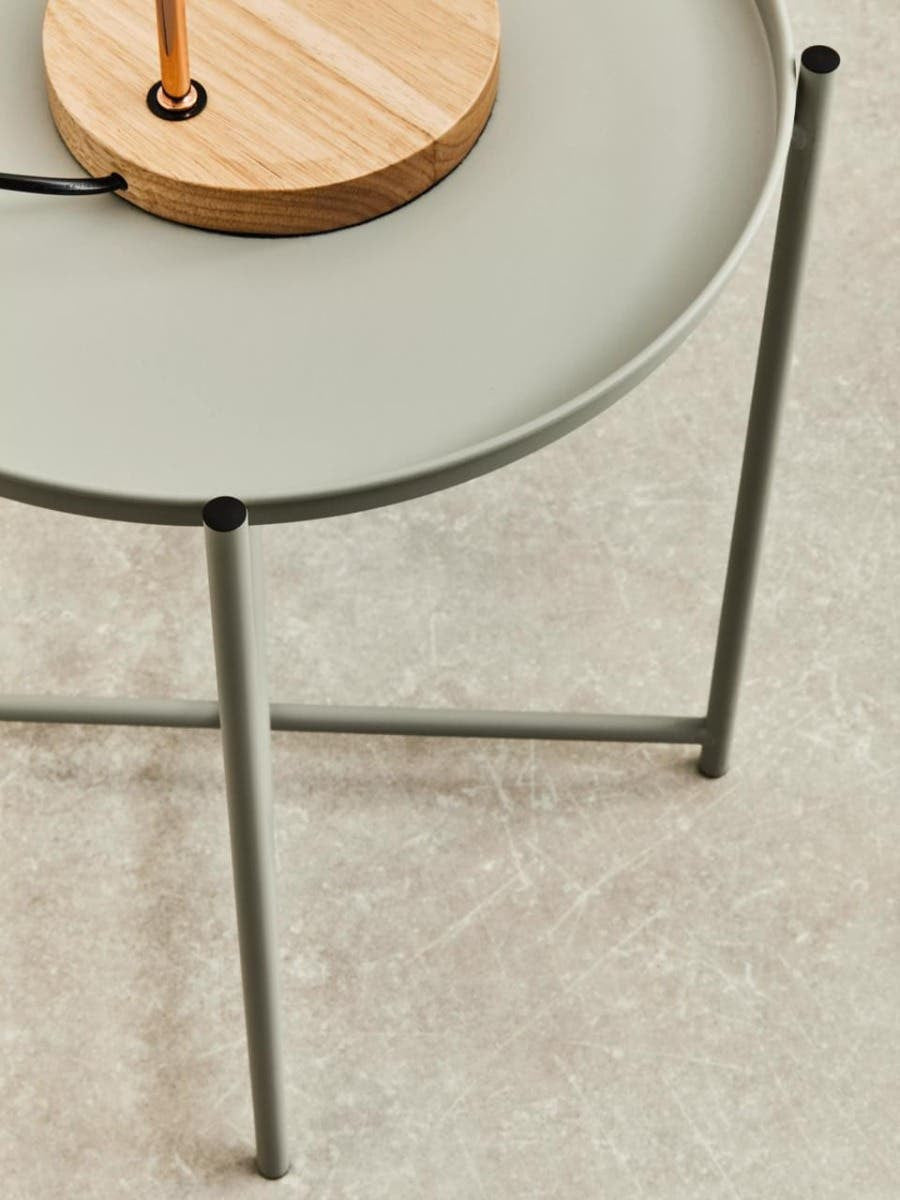 A grey iron side table with a round top and slender legs, featuring a wooden lamp base on the surface. The table is positioned on a light-colored floor, highlighting its modern design