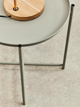 A grey iron side table with a round top and slender legs, featuring a wooden lamp base on the surface. The table is positioned on a light-colored floor, highlighting its modern design