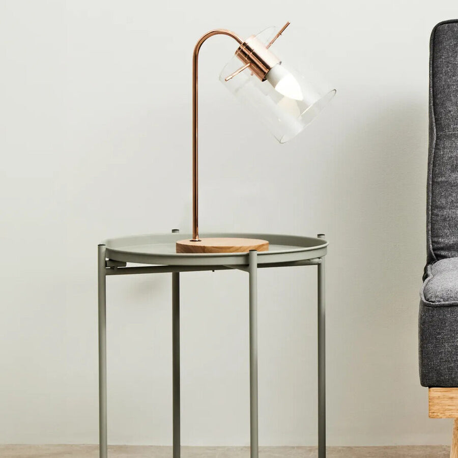A grey iron side table with a minimalist design, featuring a square top and slender legs, positioned next to a modern sofa. A stylish lamp with a curved neck and wooden base is placed on the table, casting a warm light. The background is a neutral wall, enhancing the contemporary aesthetic of the scene