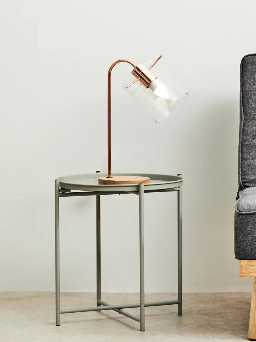 A grey iron side table with a minimalist design, featuring a square top and slender legs, positioned next to a modern sofa. A stylish lamp with a curved neck and wooden base is placed on the table, casting a warm light. The background is a neutral wall, enhancing the contemporary aesthetic of the scene
