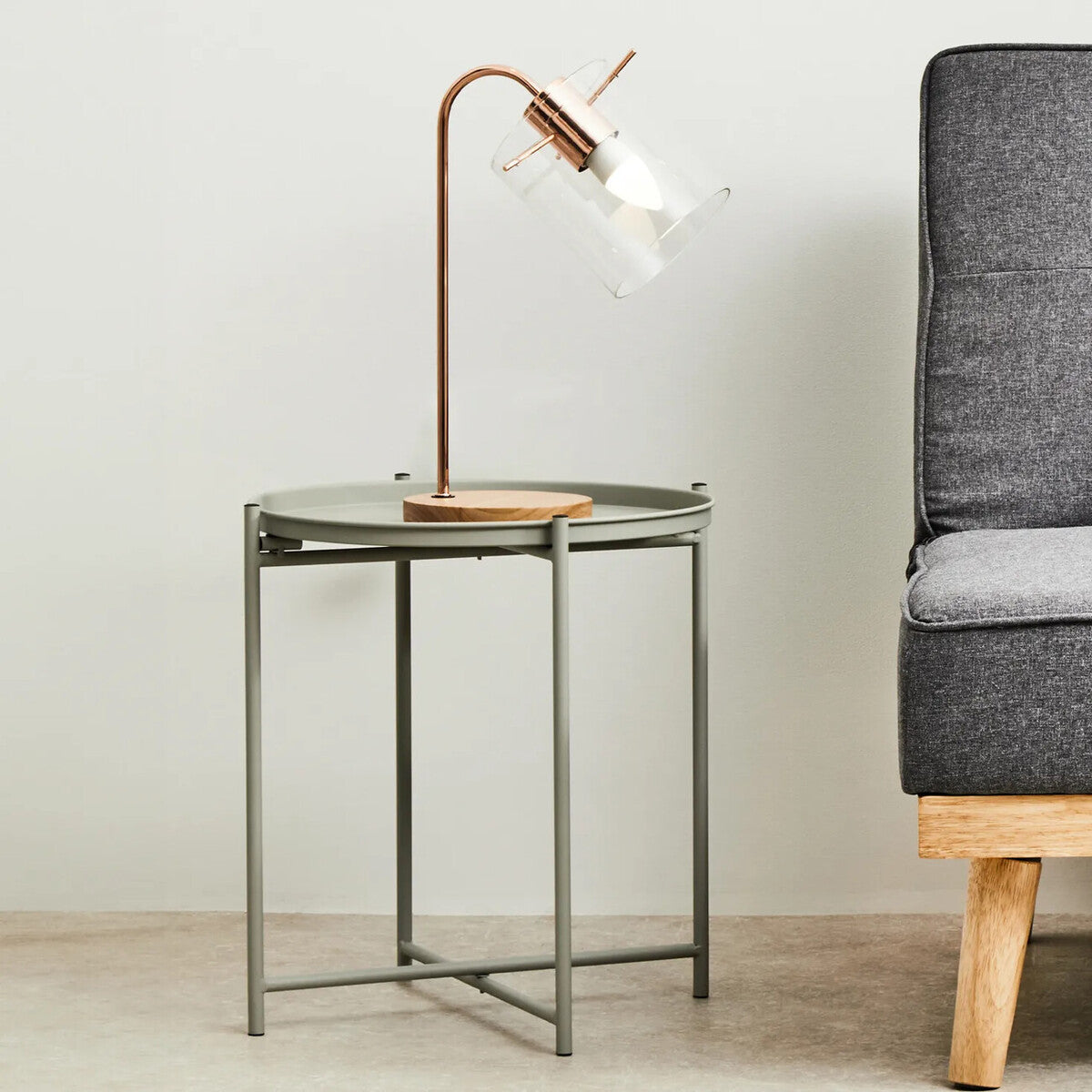 Grey iron side table with a sleek, modern design featuring a glass top and a geometric base, accompanied by a stylish lamp with a curved neck and a warm light, positioned next to a grey upholstered sofa on a light-colored floor